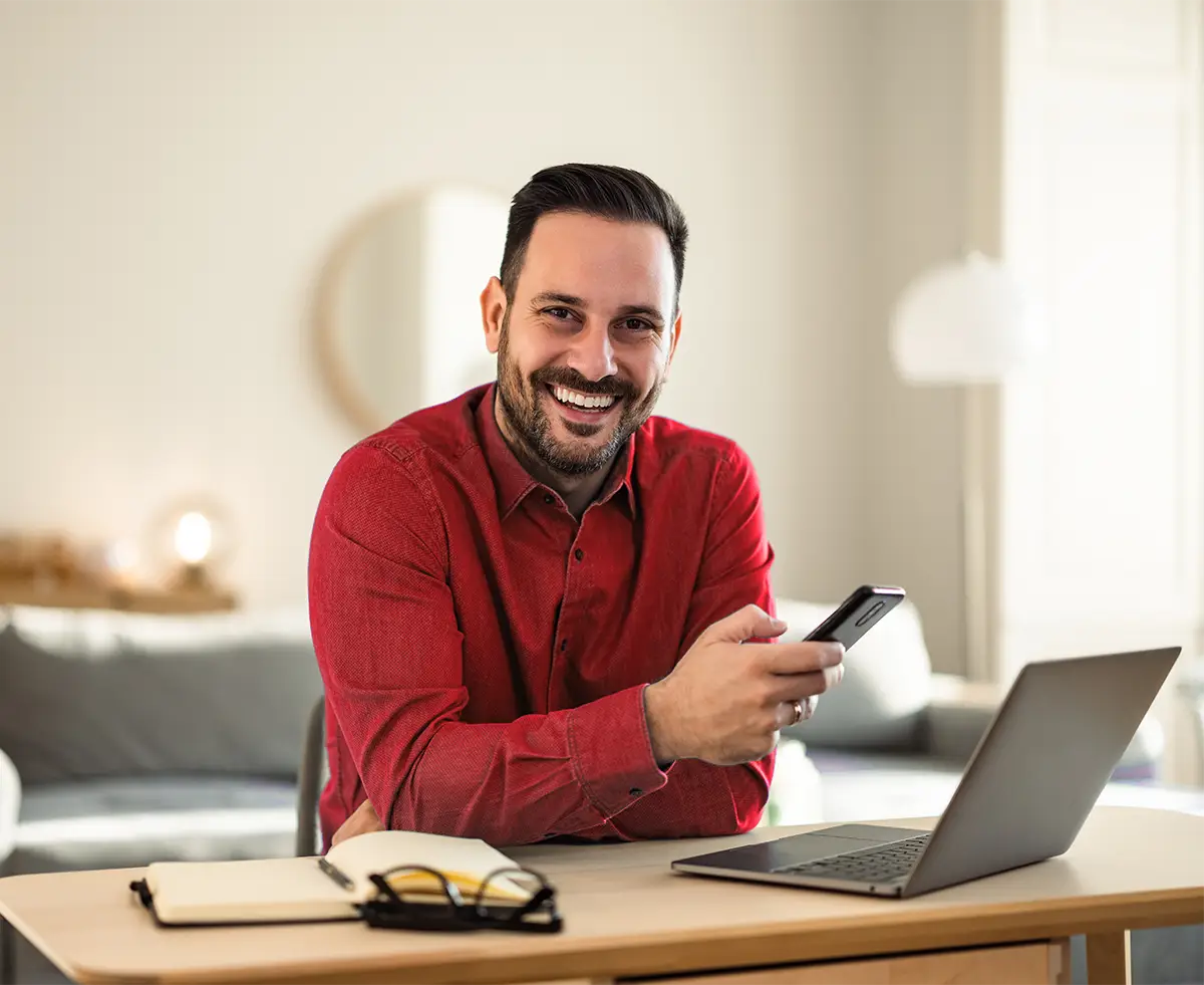 Ein lächelnder Mann in rotem Hemd sitzt an einem Schreibtisch mit Laptop, Smartphone und Notizbuch in einem hellen, modernen Wohnzimmer.